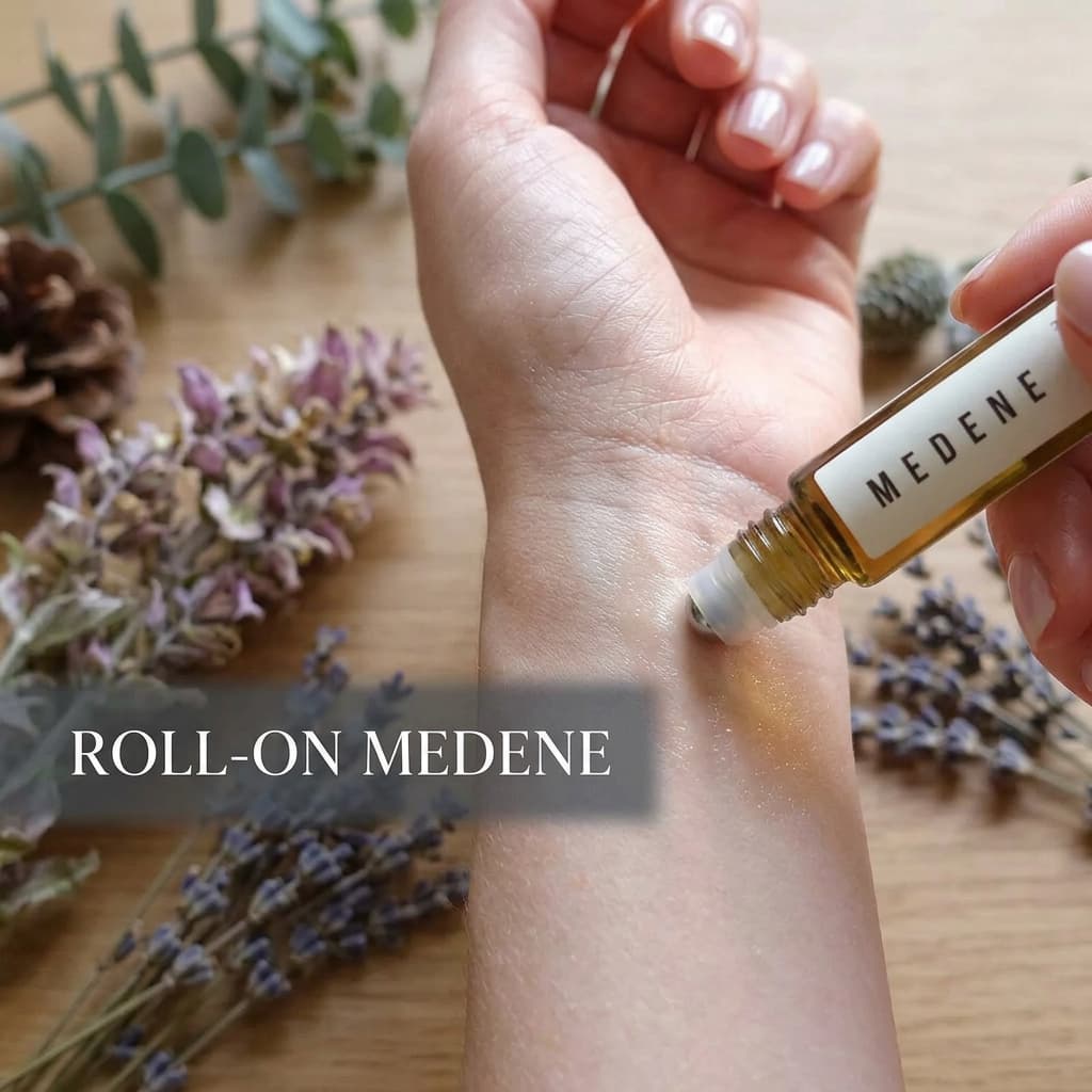 Flacon roll-on Medene appliqué sur le poignet d'une femme à la peau satinée avec des brins de lavande et de sauge flous et un texte superposé ROLL-ON MEDENE.