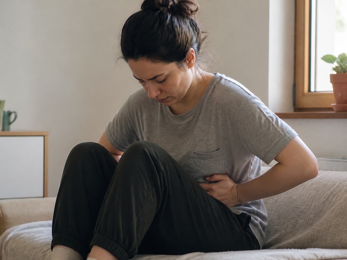 Personne assise en position repliée, mains pressant le bas-ventre, visage neutre, posture de soulagement.